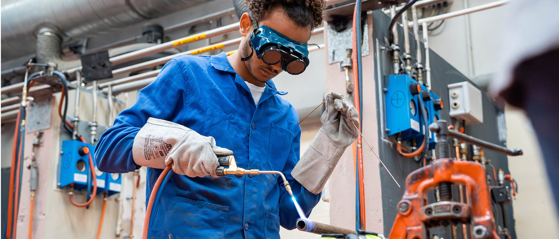 un homme dans un atelier technique tient un appareil à soudure. Il est vêtu d’une combinaison bleue, gants et lunettes de protection