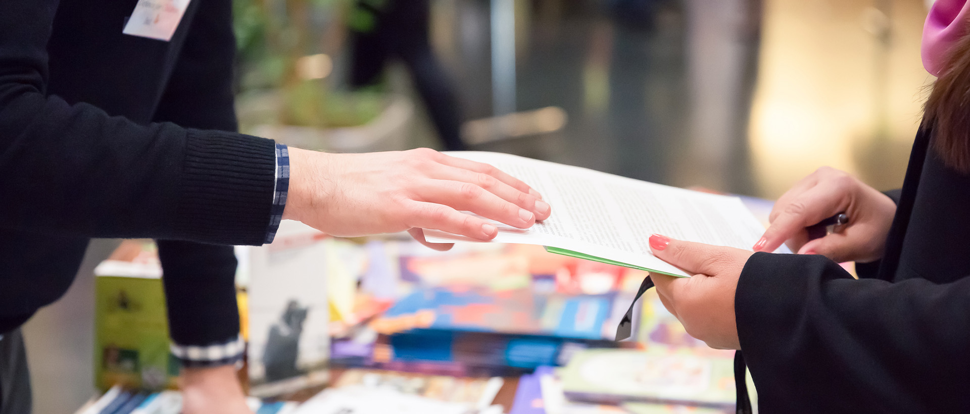 Texte alternatif : Au premier plan, deux mains s’échangent un document, au-dessus d’une table remplie de flyer, au second plan