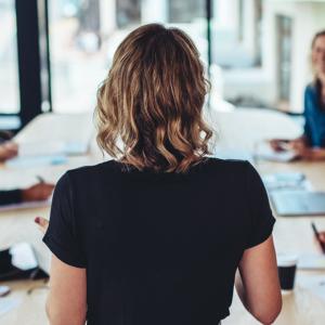 Une femme se tient devant 5 personnes, assises à une table de réunion