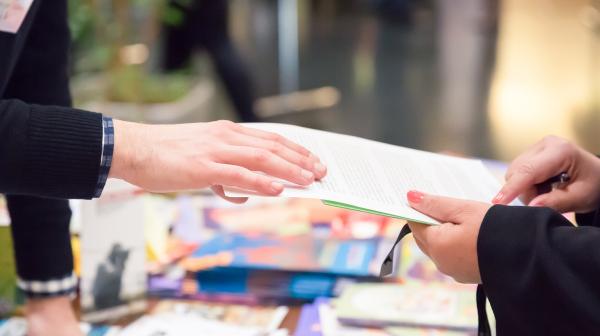 Texte alternatif : Au premier plan, deux mains s’échangent un document, au-dessus d’une table remplie de flyer, au second plan